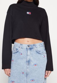 Black cropped sweatshirt with a high neckline, featuring a small multicolored "Tommy Jeans" logo; paired with a light blue denim skirt adorned with circular logos.