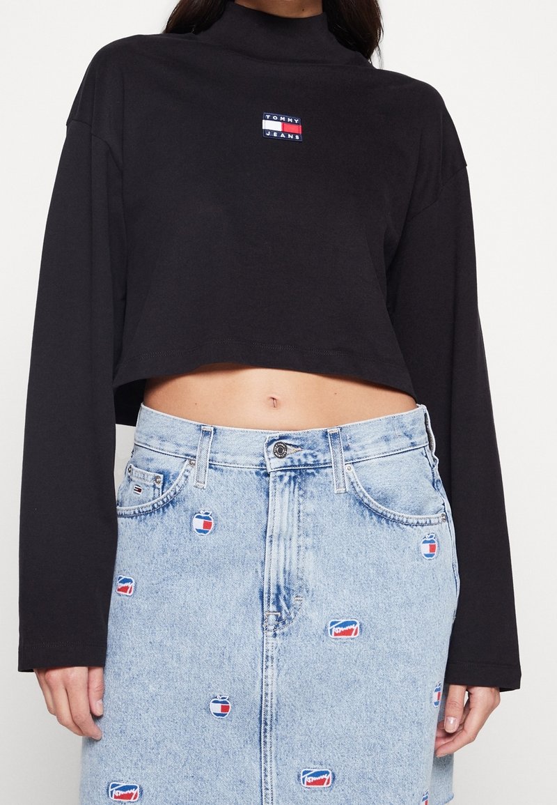 Black cropped sweatshirt with a high neckline, featuring a small multicolored "Tommy Jeans" logo; paired with a light blue denim skirt adorned with circular logos.