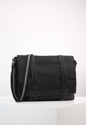 Sac messager noir avec une bandoulière réglable présentant une bande grise, exposé sur un piédestal blanc sur un fond uni.