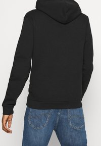 Sudadera con capucha negra, hecha de una mezcla de algodón, que presenta un bolsillo tipo canguro, puños acanalados y un corte relajado. Combinada con jeans de mezclilla azules.