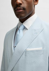 Ljusblå texturerad kavaj med notch-lapp, i kombination med en ljusblå mönstrad slips och en vit fickduk. Skräddarsydd passform.