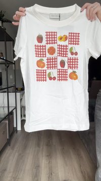 Camiseta de algodón blanca con un diseño de estampado de frutas sobre un patchwork de cuadrados a cuadros rojos y blancos. Manga corta y cuello redondo.