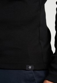 Gros plan sur l'ourlet d'une chemise noire à manches longues avec une petite étiquette noire comportant des lettres blanches entrelacées sur un pantalon bleu marine.