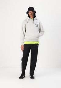 Lys grå hettegenser med lommer foran og logo, lagd over en lys grønn skjorte. Svart bøttehatt, svarte bukser og svarte joggesko med hvite striper.
