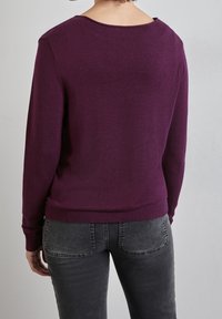 Person som bär en långärmad lila tröja och blekta svarta jeans, visad bakifrån mot en enkel grå bakgrund.