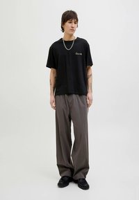 Czarny bawełniany t-shirt z detalem z logo, zestawiony z luźnymi szarymi spodniami. Model ma na sobie masywne czarne buty, stoi na neutralnym tle.