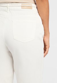 Pantalon en denim blanc avec une texture lisse, présentant une poche arrière et une étiquette de marque en cuir marron au-dessus de la taille.