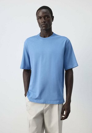 Homme portant un t-shirt bleu clair uni à manches courtes avec un pantalon beige, debout avec une main dans la poche devant un fond clair uni.