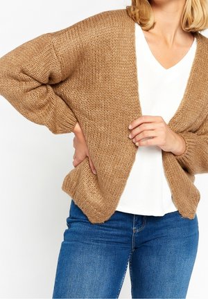 Femme portant un cardigan en grosse maille couleur chameau par-dessus un haut blanc à col en V et un jean bleu, visible des épaules aux hanches.