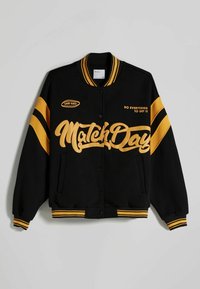 Svart varsityjacka med gula ränder och text. Har tryckknappar, ribbad krage och sidofickor. "Match Day" tryckt tydligt.