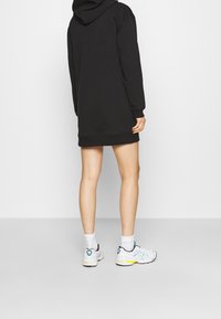 Schwarzes Kapuzenkleid mit langen Ärmeln, gerippten Bündchen und Saum. Kombiniert mit weißen Sneakers mit bunten Akzenten und kurzen Socken.