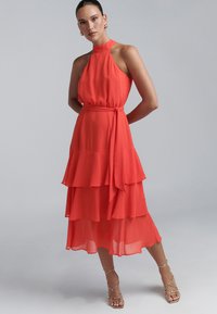 Flowy red chiffon dress with a high neck, sleeveless design, tiered ruffles, and a tied waist belt. Features a mid-calf length.