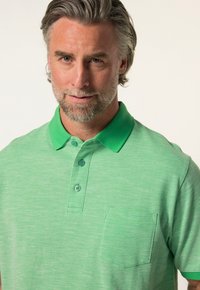 Grünes Poloshirt mit strukturierter Oberfläche, ausgestattet mit einer Knopfleiste mit zwei Knöpfen, kontrastierendem Kragen und einer Brusttasche auf der linken Seite.
