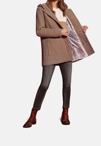 Cappotto textured marrone con cappuccio, chiusura a bottoni e grandi tasche frontali. Foderato con un leggero tessuto con motivo a logo. Indossato sopra dei jeans grigi e stivali.