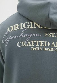 Grå hættetrøje med tekstureret skrift i creme og lyslilla. Teksten inkluderer "ORIGINAL Copenhagen," "CRAFTED," og "DAILY BASIC."