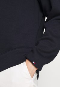Felpa navy con maniche lunghe, polsini a costine e un piccolo logo tricolore sull'orlo. Tessuto liscio, design aderente.