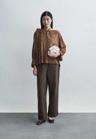 Kvinne står mot ensfarget bakgrunn iført brun semsket topp med bred krage, brune mønstrete bukser, mørke flate sko og holder en kremfarget clutchveske.