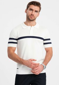 Polo shirt bianco a maniche corte realizzato in tessuto a costine, con strisce blu navy sul petto e sulle maniche. Include una chiusura a due bottoni.