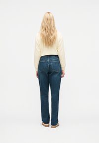 Ξανθιά γυν αίκα που στέκεται γυρισμένη με την πλάτη, φοράει κρεμ πουλόβερ, τζιν υψηλής μέσης μπλε και σουέντ παπούτσια με λευκές σόλες.