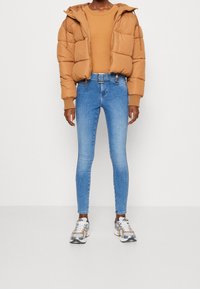Persona con una chaqueta acolchada color beige, un suéter beige a juego, jeans ajustados azul claro con cinturón y zapatillas plateadas, de pie contra un fondo liso.