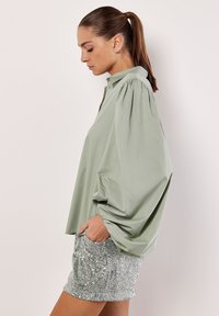 Hellgrünes Blusenshirt mit puffförmigen Ärmeln und Knopfverschluss, kombiniert mit silbernen Pailletten-Shorts mit strukturiertem Finish und Seitentaschen.
