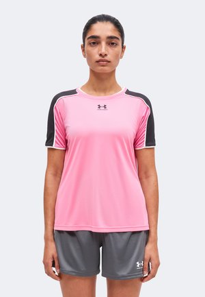 Kvinde iført en pink og sort kortærmet sportstrøje og grå atletikshorts, stående med fronten mod kameraet mod en ensfarvet baggrund.