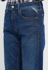 Mørkeblå denimjeans med smal passform, med oransje sømmer, en frontlomme og en logomerking på midjebåndet.