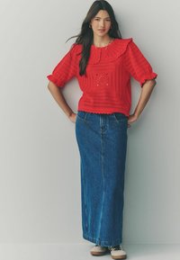Blouse en crochet rouge avec un large col et des manches courtes bouffantes, associée à une longue jupe en denim. Les chaussures sont claires avec des lacets foncés.