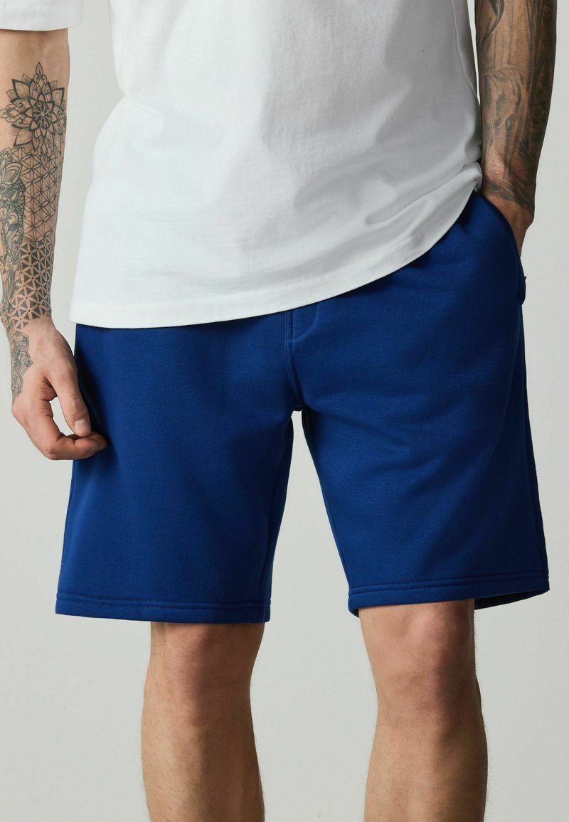 Homme portant un t-shirt blanc uni et un short décontracté bleu longueur genoux avec des bras et des mains tatoués visibles sur les côtés.