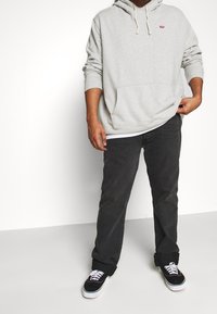 Persona che indossa un hoodie Levi's grigio chiaro, jeans neri e sneakers nere con lacci bianchi, in piedi contro uno sfondo bianco.
