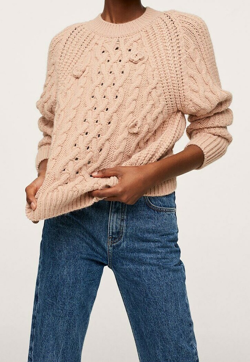 Personne portant un pull en maille épaisse beige avec des motifs tressés et un jean taille haute bleu, tenant l'ourlet du pull avec les deux mains.