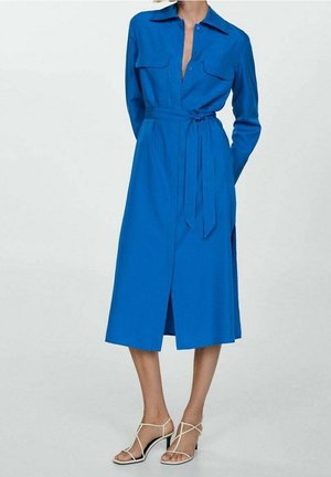 Vrouw draagt een felblauwe midi-overhemdjurk met lange mouwen en een tailleceintuur, gecombineerd met witte sandalen met hakken en bandjes, staand tegen een effen achtergrond.