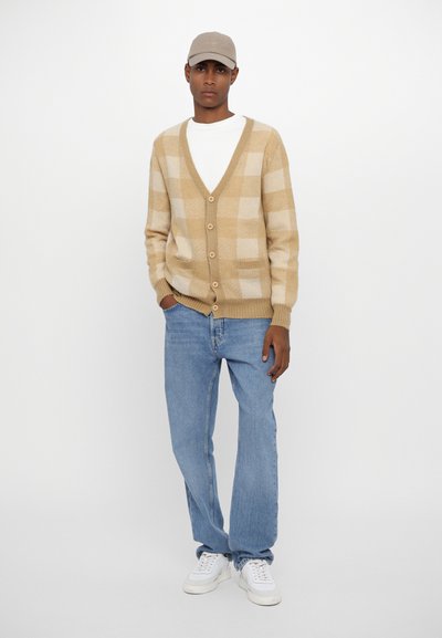 Beige- und cremefarbener Karomuster-Strickcardigan mit V-Ausschnitt, Knöpfen und Seitentaschen. Kombiniert mit hellblauen Jeans und weißen Sneakers. Eine legere Mütze getragen.