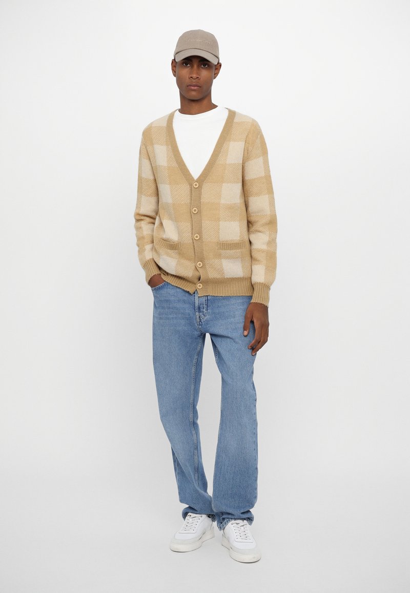 Beige och krämrandig cardigan med v-ringning, knappar och sidofickor. Kombinerad med ljusblå jeans och vita sneakers. En avslappnad keps bärs.