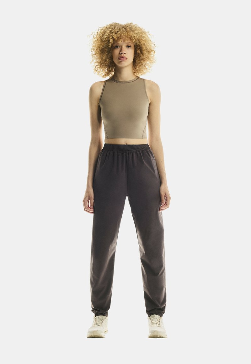 Mlada osoba s kovrčavom plavom kosom, koja nosi bezrukavi bež crop top, crne trenirke i bijele tenisice, stoji okrenuta prema naprijed.