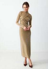 Robe longue et ajustée en tissu doré brillant avec un col montant, un design drapé sur la poitrine et des manches longues.