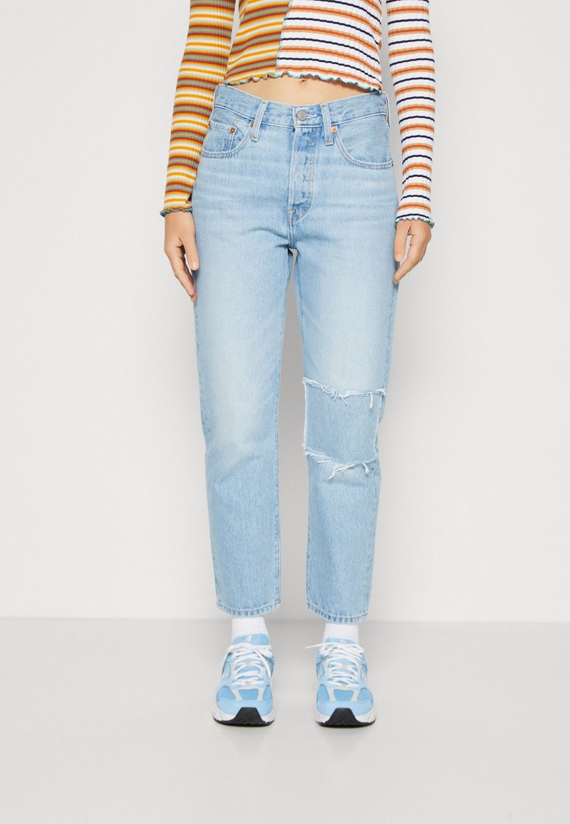 Jeans taille haute bleu clair avec détails de genoux déchirés, coupe droite et décoloration subtile. Associés à des baskets bleu clair.