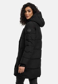 Abrigo acolchado negro con capucha, que cuenta con un diseño acolchado y un parche de logo en la manga. Llega hasta la mitad del muslo. Material suave y texturizado.