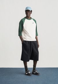 Jeune homme portant une casquette Nike bleue, un t-shirt Nike blanc et vert, un short noir, des baskets Nike noires et des chaussettes foncées, debout sur un tapis bleu contre un mur blanc.