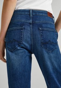 Jeans de mezclilla azul oscuro con una cintura de media altura, dos bolsillos traseros con un diseño de costura ondulada y un parche de cuero en la cinturilla.