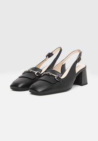 Scarpe slingback in pelle nera con punta squadrata, tacco chunky e catena metallica decorativa sulla tomaia. Texture liscia, design minimal.