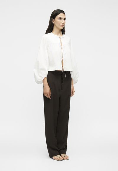 Γυναίκα με μακριά σκούρα μαλλιά που φοράει λευκό μπλουζάκι με δέσιμο μπροστά και φουσκωτά μανίκια, μαύρο παντελόνι με φαρδιά πόδια και μπεζ σανδάλια, όρθια πλάγια.