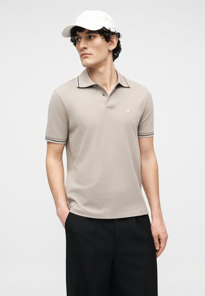 Νέος άνδρας που φοράει μπεζ πόλο μπλούζα, μαύρο παντελόνι και λευκό καπέλο, στεκόμενος με το ένα χέρι στην τσέπη, κοιτάζοντας προς τα πλάγια μπροστά σε απλό φόντο.