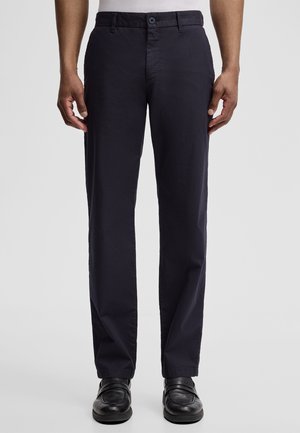 Mann trägt schwarze Hosen mit geradem Bein und schwarze Lederschuhe, steht vor einem schlichten hellen Hintergrund.