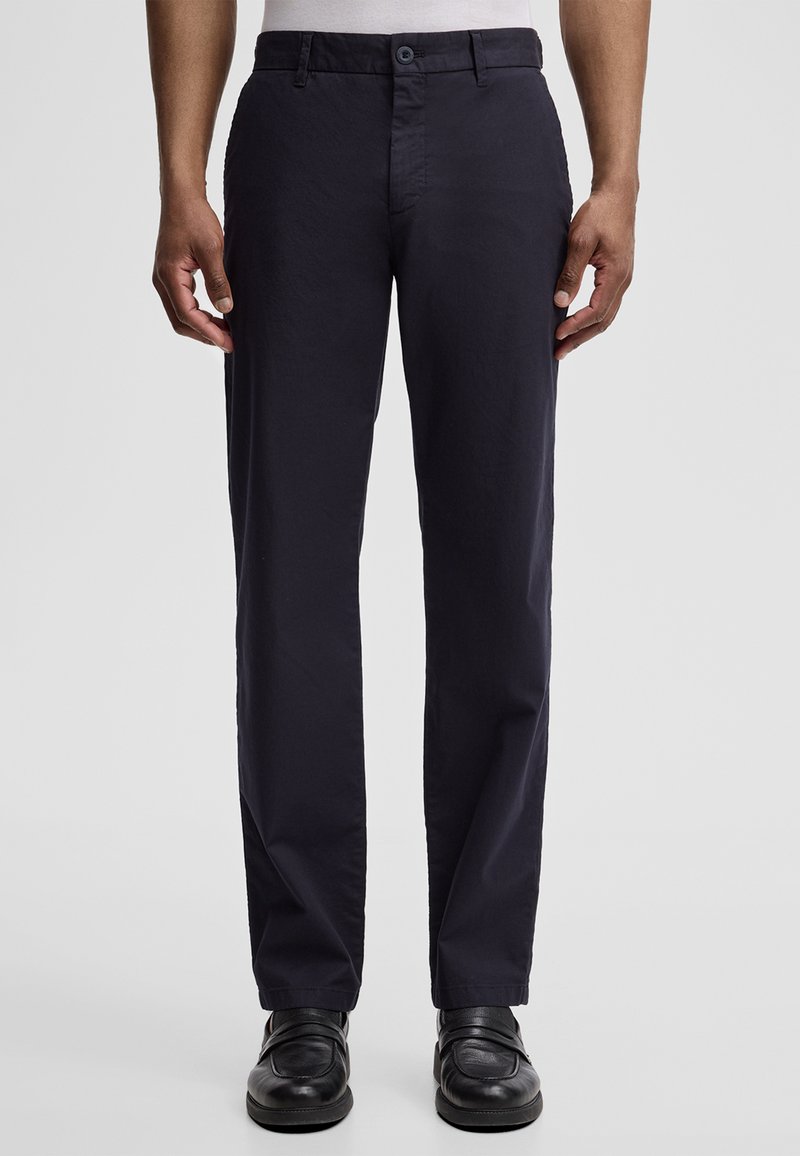 Uomo che indossa pantaloni neri a gamba dritta e scarpe di pelle nere, in piedi contro uno sfondo chiaro e semplice.