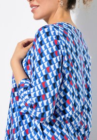 Top blu patterned con un design geometrico e accenti bianchi, rossi e neri. Tessuto leggero con maniche a tre quarti e una vestibilità rilassata.