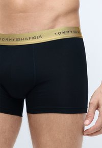 Sorte boxer shorts lavet af blødt stof med en guldbelagt talje, hvor der står "TOMMY HILFIGER" trykt med sorte bogstaver. Glat tekstur, ingen synlige mønstre.