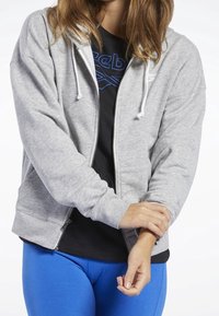 Felpa grigia con zip e cordini, dalla texture morbida, con due tasche laterali, indossata sopra una maglietta nera e abbinata a leggings blu.