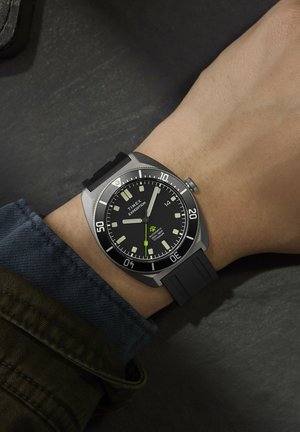 Poignet d'homme portant une montre Timex Expedition à cadran noir avec des marqueurs lumineux et un bracelet en caoutchouc noir sur une surface sombre.
