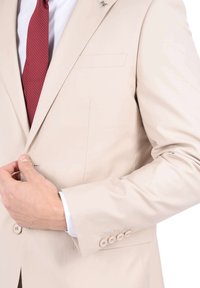 Giacca di completo beige chiaro con una texture liscia, revers a punta, fazzoletto da taschino e bottoni decorativi sulle maniche. Completata da una cravatta a pois rossi.